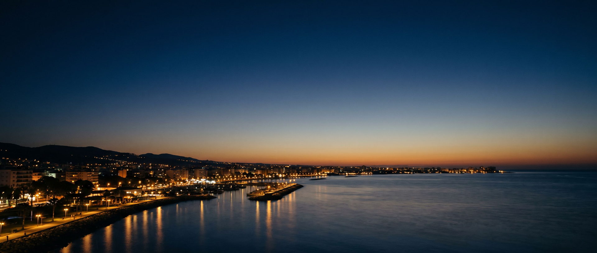 Die Küste von Limassol zur blauen Stunde: warme Lichter der Uferpromenade und Marina spiegeln sich im ruhigen Mittelmeer unter tiefblauem Abendhimmel